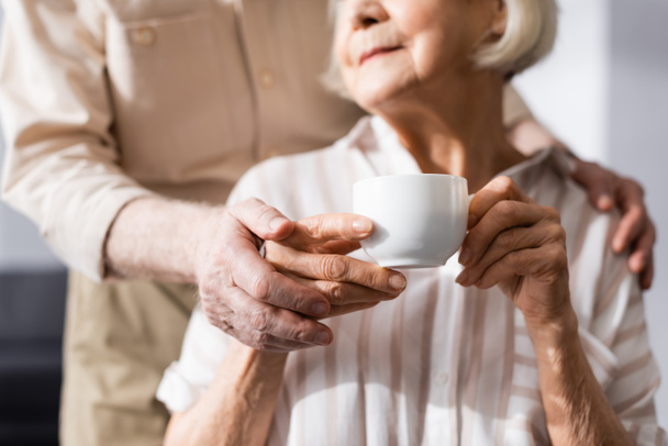 Cropped view of senior man embracing wife holding coffee cup at home  - Foto, Bild
