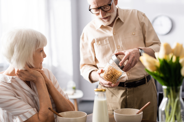 Szelektív fókusz idős férfi nyitó üveg gabonafélék közelében feleség reggeli közben a konyhában  - Fotó, kép