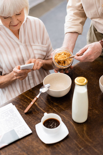 Enfoque selectivo del señor hombre vertiendo cereales cerca de la esposa utilizando el teléfono inteligente en la mesa
  - Foto, Imagen