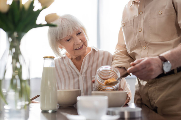 Selektivní zaměření usměvavé starší ženy sedí v blízkosti muže nalévání cereálií v misce během snídaně  - Fotografie, Obrázek