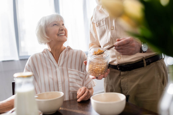 Selektywne skupienie starszej kobiety uśmiechającej się do męża wylewającego płatki ze słoika podczas śniadania  - Zdjęcie, obraz