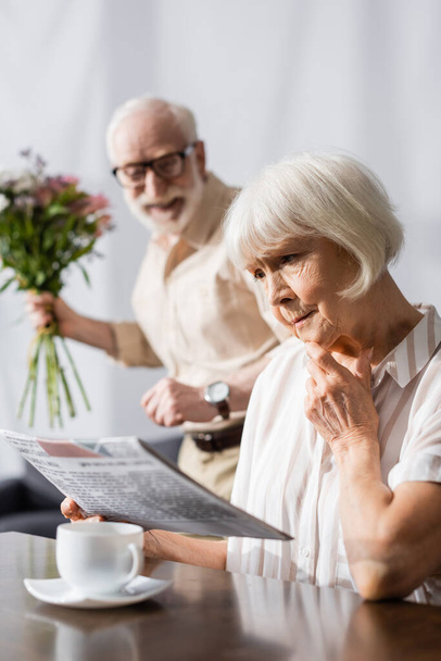 Selective focus of sad senior woman reading news near positive husband with bouquet at home  - Photo, Image