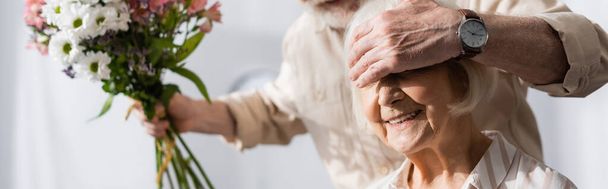 Πανοραμική φωτογραφία του ηλικιωμένου άνδρα που καλύπτει τα μάτια σε χαμογελαστή σύζυγο και κρατώντας λουλούδια μπουκέτο στο σπίτι - Φωτογραφία, εικόνα
