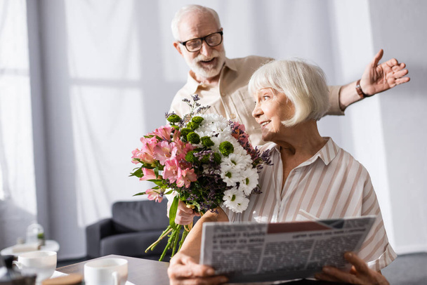 Selective focus of positive senior man holding bouquet near wife with newspaper in kitchen  - Foto, afbeelding