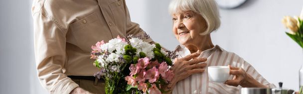 Panoramic shot of senior man giving bouquet to smiling wife with cup of coffee  - Foto, Imagem