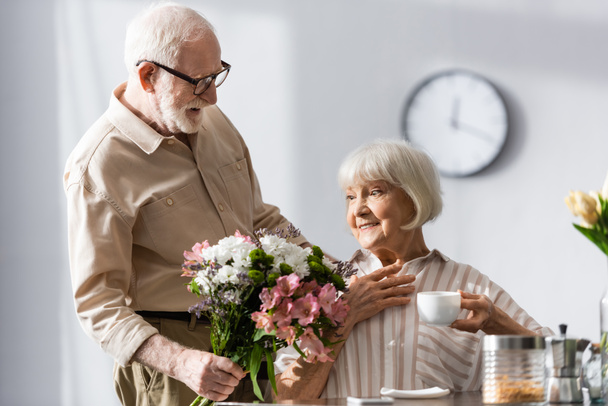 Selective focus of elderly man giving bouquet to positive wife with cup of coffee at home  - Fotó, kép