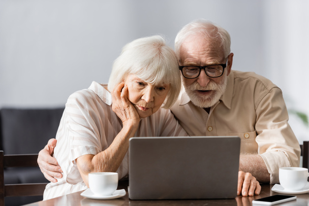 Selektywne skupienie starszego mężczyzny obejmującego żonę podczas korzystania z laptopa w pobliżu filiżanek kawy i smartfona na stole  - Zdjęcie, obraz