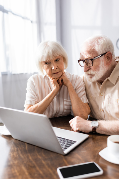 Foco seletivo de casal de idosos olhando para laptop perto de smartphone e café na mesa
  - Foto, Imagem