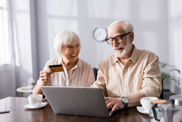 Valikoiva painopiste hymyilevä vanhempi pari käyttää kannettavaa tietokonetta ja luottokorttia lähellä kahvikupit pöydällä  - Valokuva, kuva