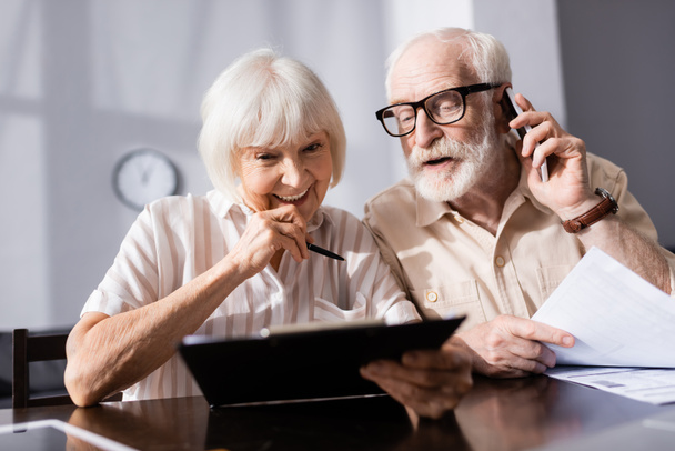 Selective focus of smiling senior woman holding clipboard near husband talking on smartphone at home   - Photo, Image