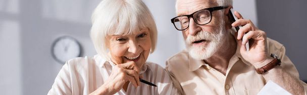 Panoramic shot of senior man talking on smartphone near wife holding pen  - Photo, Image