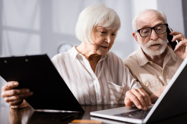 Selective focus of senior woman holding clipboard and using laptop near husband talking on smartphone   - Fotografie, Obrázek
