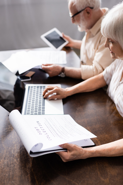 Selective focus of senior woman holding papers with debt collection lettering and using laptop near husband at home  - Photo, Image