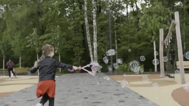Active little blond girl with a braid creating bubbles while running at the city park. Action. Adorable excited female child playing with soap bubbles at the playground, rear view. - Video, Çekim
