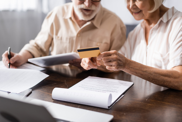 Focus selettivo della donna anziana in possesso di carta di credito mentre il marito scrive su carte vicino gadget sul tavolo  - Foto, immagini