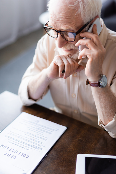 Senior άνθρωπος μιλάει στο smartphone κοντά σε χαρτιά με επιστολές συλλογής χρέους και ψηφιακή ταμπλέτα στο τραπέζι  - Φωτογραφία, εικόνα
