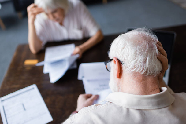 Selective focus of elderly man with hand near head holding papers near wife at home  - Foto, Imagem
