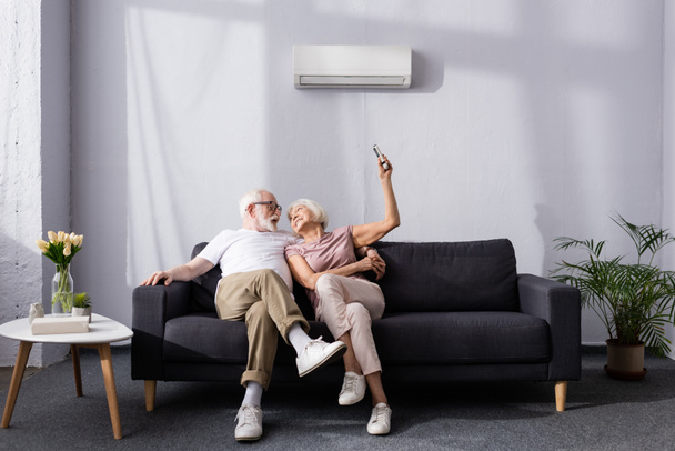Χαρούμενο ζευγάρι ηλικιωμένων που χρησιμοποιούν τηλεχειριστήριο του κλιματιστικού στον καναπέ  - Φωτογραφία, εικόνα