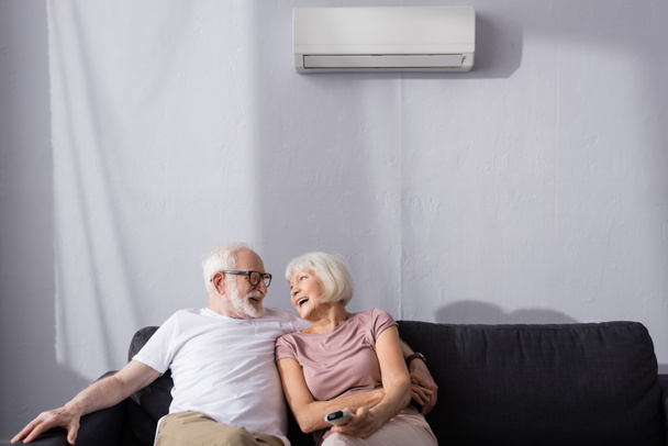 Senior man embracing positive wife with remote controller of air conditioner on couch  - Foto, imagen