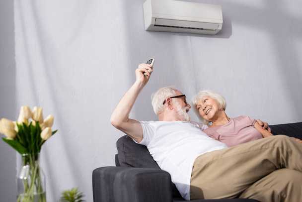 Selective focus of smiling man switching air conditioner with remote controller near wife  - Photo, Image