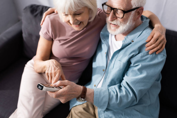 Enfoque selectivo de la sonriente pareja de ancianos utilizando el controlador remoto de aire acondicionado en el hogar
  - Foto, imagen