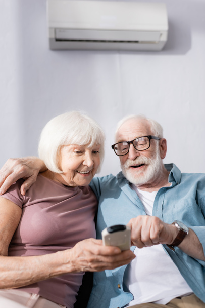Enfoque selectivo de sonreír al hombre mayor abrazando a la esposa y utilizando el controlador remoto de aire acondicionado en el hogar
   - Foto, imagen