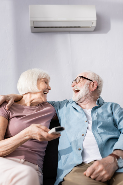 Enfoque selectivo de reír hombre mayor abrazando esposa con controlador remoto de aire acondicionado
  - Foto, Imagen
