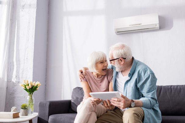 Hombre mayor sosteniendo la tableta digital y abrazando a la esposa sonriente en el sofá
  - Foto, imagen