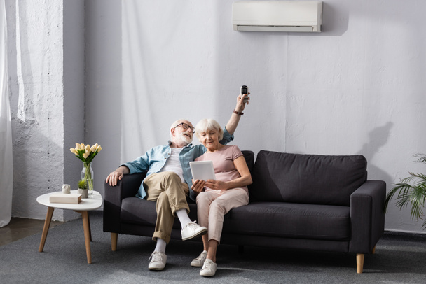 Donna anziana che utilizza tablet digitale vicino sorridente marito commutazione condizionatore d'aria con telecomando  - Foto, immagini