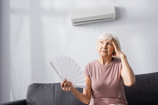 Donna anziana stanca con mano vicino alla testa che tiene ventilatore mentre si sente caldo a casa  - Foto, immagini