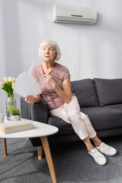 Επιλεκτική εστίαση της ηλικιωμένης γυναίκας κρατώντας ανεμιστήρα, ενώ αίσθημα θερμότητας στο σαλόνι - Φωτογραφία, εικόνα