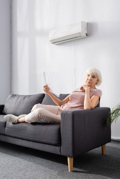 Ältere Frau wedelt mit Ventilator unter Klimaanlage im Wohnzimmer - Foto, Bild