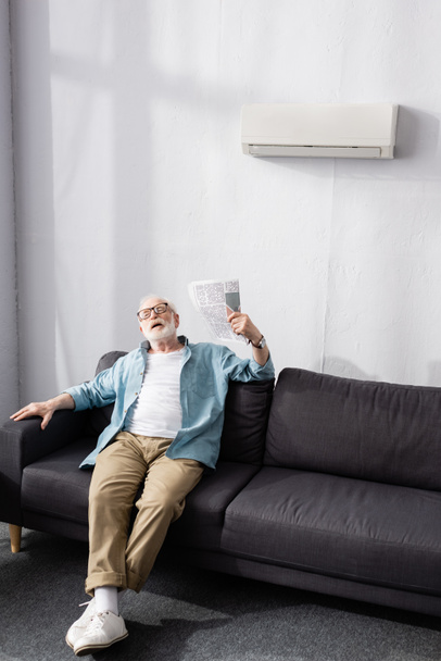 Senior man waving newspaper while suffering from heat on couch  - Фото, зображення