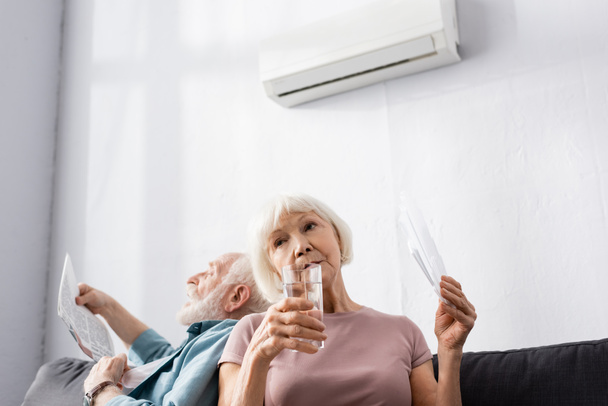Selective focus of elderly woman holding glass of water and fan near husband suffering from heat at home  - Fotografie, Obrázek