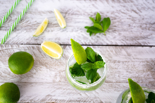 One glass with a refreshing drink with ice, mint leaves and a slice of lime on a light wooden background. Near the glass are three slices of juicy lime, fresh mint leaves and cocktail tubes, top view. - Фото, изображение