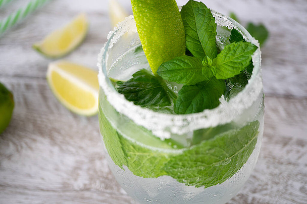 Mojito refreshing cocktail drink with ice, lime and mint in a glass on a light wooden background. Lime slices lie near. The edges of the glass are grated with lime and covered with salt, close-up. - Foto, Imagen