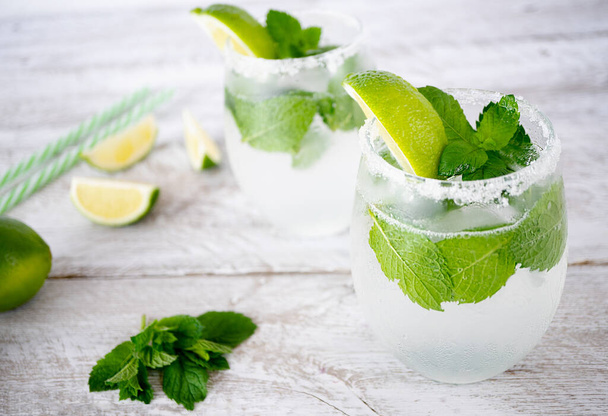 Two glasses with cool drinks with ice, lime and mint stand on a light wooden background. The edges of the glasses are coated with sugar. Nearby are fresh mint leaves and lime wedges. - Foto, Imagen