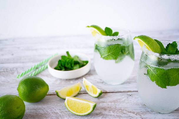 Two glasses with summer cold drinks with lime and mint stand on a light wooden table. Lime slices lie near and there is a white bowl with mint. The edges of the glasses are coated with sugar. - Foto, Imagen