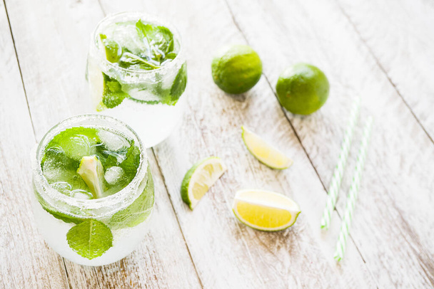 Fresh cool drinks with lime, mint and ice in glasses stand on a light wooden background. Near by lie slices of lime and cocktail tubes. The concept of a healthy diet, fitness. - Foto, Imagen