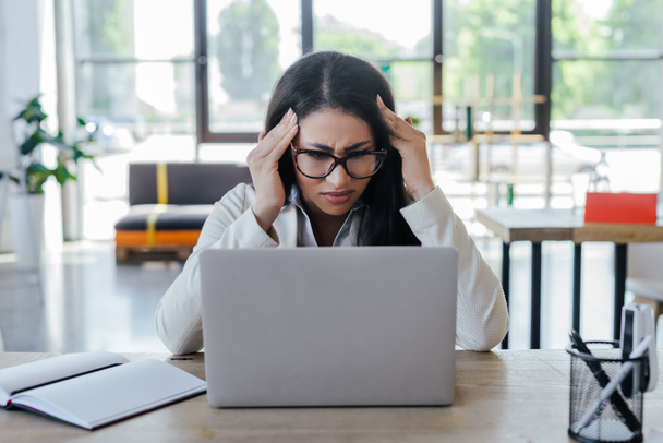 stressed businesswoman in glasses using laptop near notebook on table - Fotoğraf, Görsel