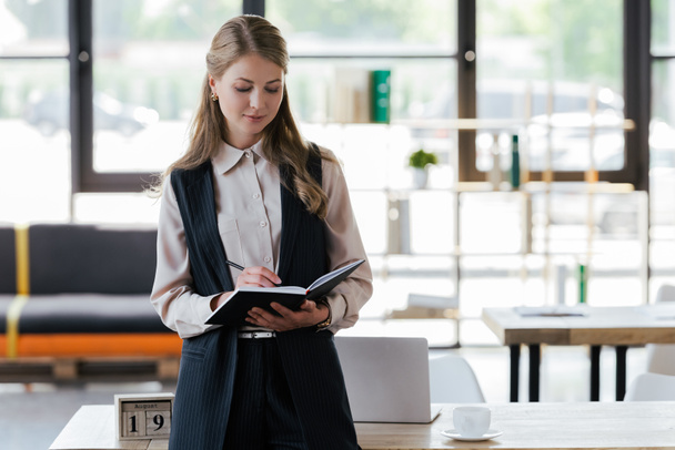 attractive businesswoman standing near table writing in notebook - Photo, image