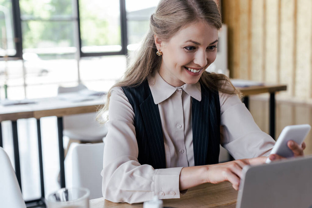 selective focus of beautiful businesswoman smiling while using smartphone in office - Zdjęcie, obraz