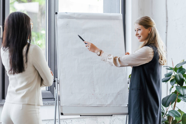 attractive businesswoman holding marker pen near flipchart and looking at coworker  - Photo, Image