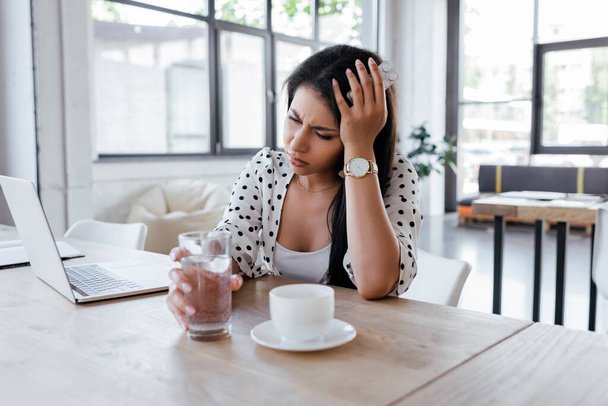 Başı ağrıyan bir kadın elinde bir bardak su, ofisinde bir fincan kahve ve dizüstü bilgisayar tutuyor. - Fotoğraf, Görsel