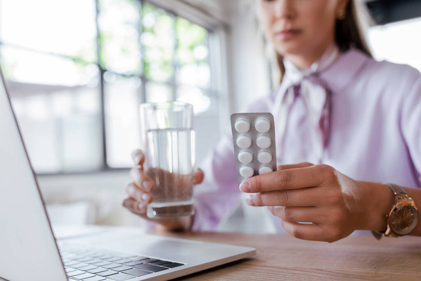 cropped view of woman holding glass of water and blister pack with pills near laptop  - Valokuva, kuva