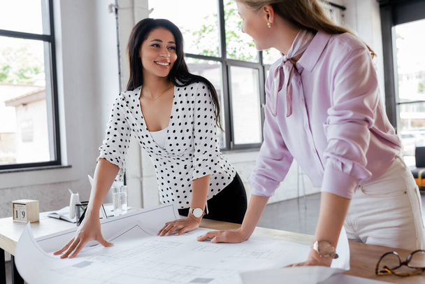 enfoque selectivo de las mujeres de negocios felices mirándose entre sí cerca de planos en la oficina
 - Foto, Imagen