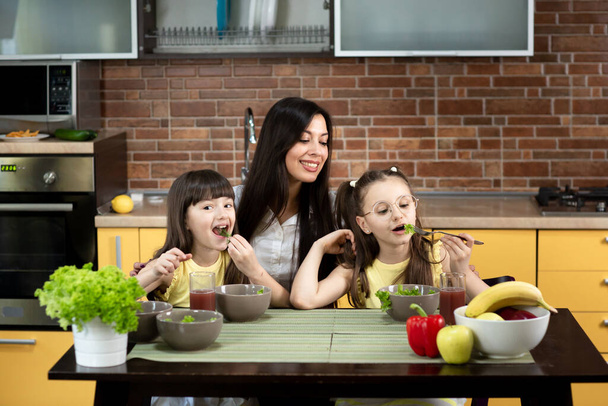 Iloinen äiti ja kaksi tytärtä syövät terveellistä salaattia yhdessä kotona. Terveellisen ruokavalion käsite, perhearvot, aika yhdessä - Valokuva, kuva