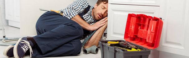Panoramic shot of plumber sleeping near pipes and toolbox in kitchen  - Foto, Imagem