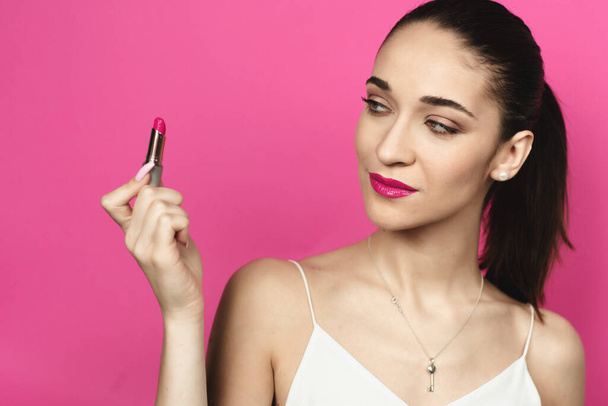 Caucasian woman with a ponytail on a pink background. Young woman with pink lips looking at lipstick. - Photo, image
