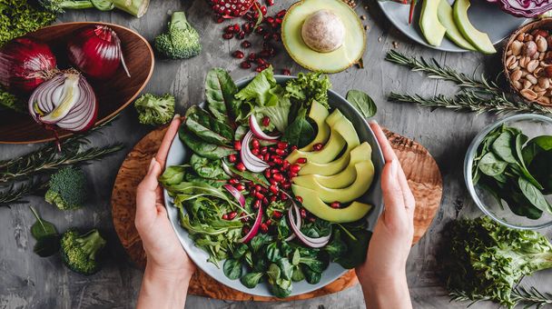 Женщина готовит здоровый свежий салат с авокадо, зелень, руккола, шпинат в тарелке на сером фоне. Здоровое веганское питание, чистое питание, диета, вид сверху, тонизация
 - Фото, изображение